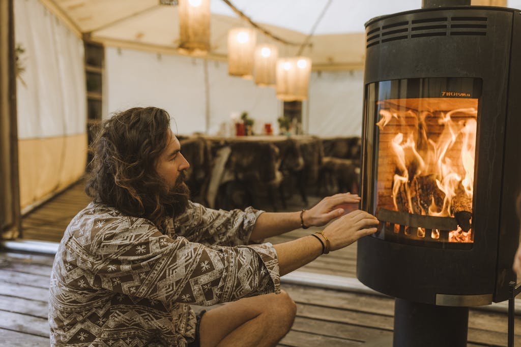 A man with long hair warming his hands by a modern stove in a cozy, well-lit tent setting.